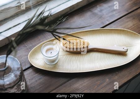 Details des Innenraums, aromatische Kerze aus Glas und Holzbürste mit natürlichen Borsten auf Metallschale auf hölzerner Fensterbank Stockfoto