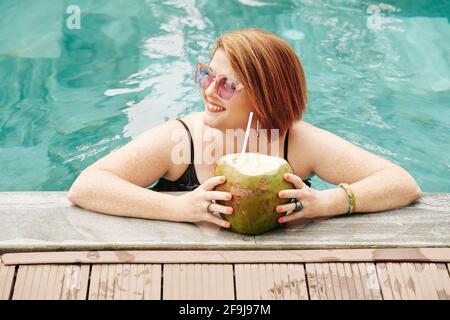 Glückliche reife Frau mit Kokosnusscocktail im Schwimmbad Und wegschauen Stockfoto