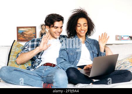 Ein glückliches junges, multirassisches Paar verbringt seine Freizeit zu Hause auf dem Sofa, mit einem Laptop, sie sprechen per Videokonferenz mit Familie oder Freunden, grüßen den Gesprächspartner, lächeln, glücklich zusammen Stockfoto