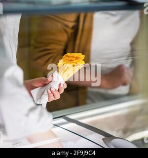 Close-up Hände des Verkäufers mit Obst Eis orange Farbe in Waffelkegel, verschwommener Hintergrund des Käufers. Echte Szene im Laden Stockfoto