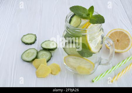 Auf dem weißen Holzhintergrund verdünntes Wasser oder Wasser mit Zitrone, Gurke und Ingwer im Glas aufgießen. Nahaufnahme. Stockfoto