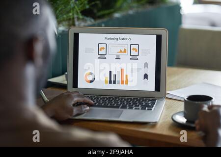 Rückansicht eines modernen afroamerikanischen Mannes, der einen Laptop mit Grafiken und Diagrammen benutzte, während er am Kaffeetisch im umweltfreundlichen Innenraum arbeitete, Kopierraum Stockfoto
