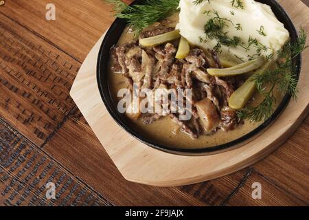 Rindfleisch Stroganoff mit Kartoffelbrei und eingelegten Gurken in einer Pfanne. Holzhintergrund Stockfoto