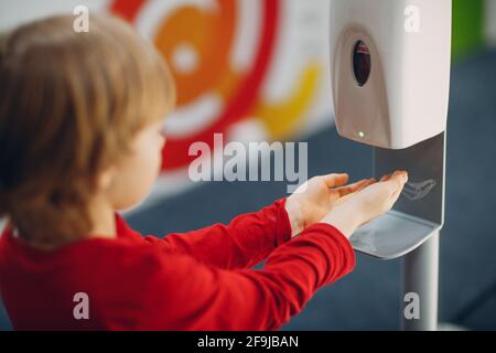 Kind Junge Kind mit automatischen Alkohol-Gel-Spender Sprühen auf Hände Desinfektionsmaschine antiseptisches Desinfektionsmittel, neues normales Leben nach Coronavirus COVID-19 Pandemie. Stockfoto