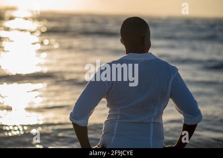 Frau, die während des Sonnenuntergangs mit reflektierendem Licht auf das Meer blickt Vom Wasser Stockfoto