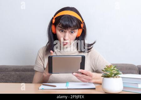 Überraschte schöne kaukasische Frau in orangefarbenen Kopfhörern, die online mit Freunden auf dem Tablet spricht, während sie zu Hause am Tisch sitzt. Bloggerin c Stockfoto