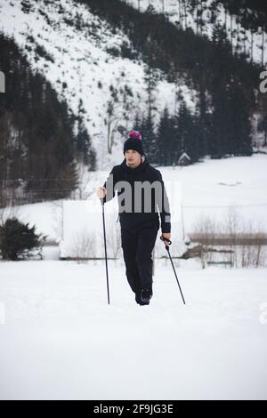Aufnahme eines Mannes in Wintersportbekleidung und eines Hutes mit Pompon, der mit Trekkingstöcken auf den Gipfel eines verschneiten Berges steigt. Echtes Porträt eines Jungen Stockfoto