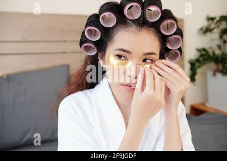 Schöne junge asiatische Frau mit Haarrollen Anwendung Patches unter Ihre Augen Stockfoto