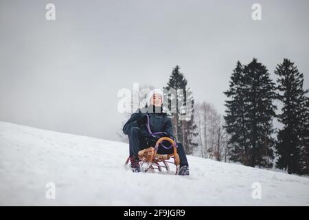 Fröhliches Lächeln eines 16-jährigen Jungen, der auf einem historischen Holzschlitten auf einer Skipiste reitet. Gefährliches Fahren. Genießen Sie den gefallenen Schnee im Winter. Narr Stockfoto