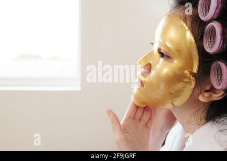 Junge Frau mit Haarrollen Anwendung Anti-Aging goldene Bettmaske Morgens auf ihrem Gesicht Stockfoto