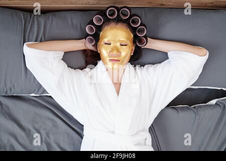 Junge Frau im Bademantel entspannt auf dem Bett mit Haarwalzen und Bettmaske auf, Blick von oben Stockfoto