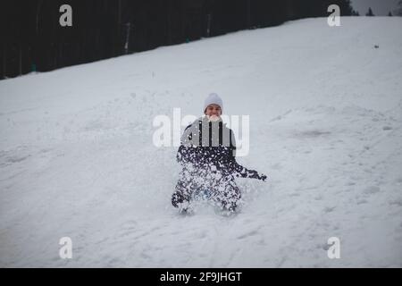 Teenager mit einem glücklichen Lächeln auf seinem Gesicht läuft auf einem Plastikboard die Pisten hinunter und stürzt sich mit hoher Geschwindigkeit hinunter. Während des Bremsens sprüht ein Schneesturm Stockfoto