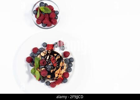 Draufsicht auf Pfannkuchen mit Schokoladensirup, Erdbeeren, Himbeeren, Heidelbeeren und Minzblättern auf weißem Teller auf weißem Hintergrund. Teller mit Scheibe Stockfoto