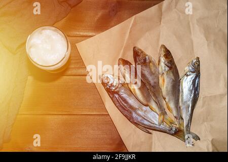 Ein Bierglas mit dunklem oder hellem geschäumtem Bier und vier gesalzenen und getrockneten, ruckartigen Fischen kaspische Kakerlake auf Bastelpapier liegen auf einem Holztisch mit braunem Brett, oben Stockfoto