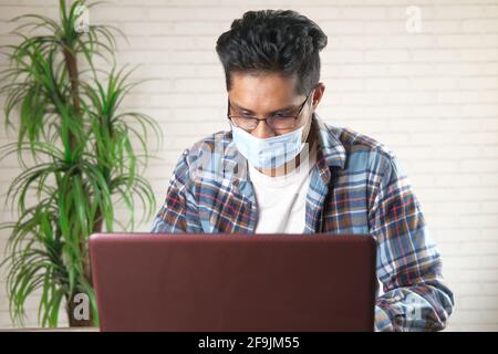 Junge asiatische Mann in Gesichtsmaske arbeiten auf Laptop Stockfoto