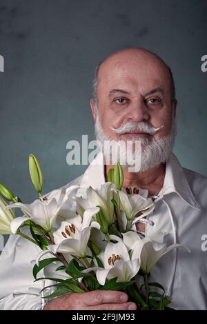 Porträt eines älteren Mannes, kahl, mit grauem Bart und lateinischer Erscheinung, trägt ein weißes Hemd und hält einen Strauß weißer Lilien Stockfoto