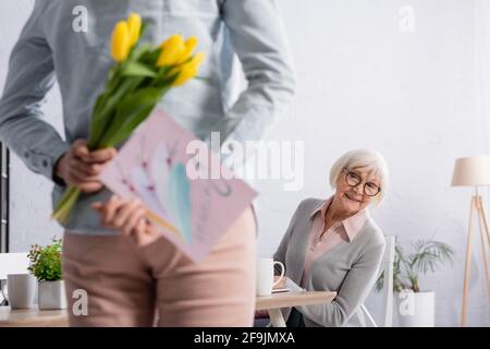 Lächelnde ältere Frau, die neben der Tochter sitzt und Blumen hält und grüßt Karte auf verschwommenem Vordergrund Stockfoto