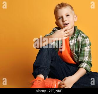 Schuljunge Teenager in kariertem Hemd, orangefarbenem T-Shirt und Jeans sitzt auf dem Boden Gespräche und Gesten Stockfoto