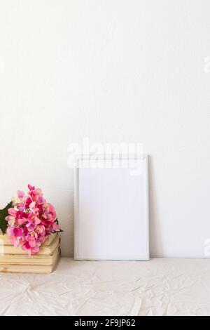 Heimdekor-Modell, leerer Bilderrahmen in der Nähe einer weiß gestrichenen Betonwand, ein Zweig rosa Hortensien auf alten Büchern und eine Kerze Stockfoto