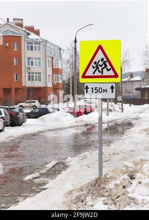 Verkehrszeichen, seien Sie vorsichtig Kinder. Schneeverwehungen mit schmutzigem Schnee und großem Wasser. Vor dem Hintergrund von Gebäuden und Autos. Menschen laufen auf weißem Hintergrund in einem roten Dreieck und einem gelben Quadrat. Stockfoto