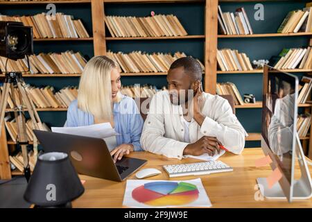 Glückliche, multiethnische Kollegen diskutieren über ein Projekt, sprechen über verschiedene Ideen, lächeln. Erfolgreiche Mitarbeiter, die ein Notebook verwenden und Papierkram machen Stockfoto