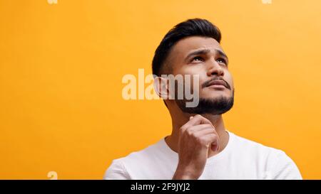 Kopf- und Schulteransicht eines nachdenklichen Mannes mit gemischter Rasse, der nach oben schaute. Erwachsener bärtiger männlicher Studioportrait, oranger Hintergrund. Stockfoto