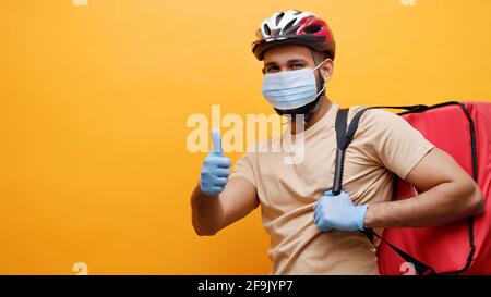 Studioaufnahme des Fahrradlieferers mit Schutzmaske und blauen Schutzhandschuhen. Blick auf die Kamera und Geste mit Daumen nach oben. Stockfoto