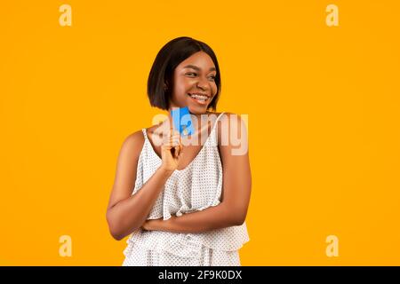Schöne schwarze Frau in stilvollem Kleid hält Kreditkarte auf Orangefarbener Studiohintergrund Stockfoto