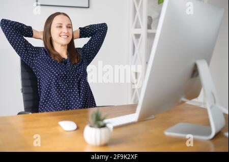 Lächelnde weibliche Mitarbeiterin macht am Arbeitsplatz im Büro eine Pause, lehnt sich in den Stuhl zurück und ist zufrieden mit dem Gesichtsausdruck, Frau freut sich über das fertige Projekt oder den Arbeitstag Stockfoto