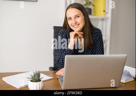 Die Unternehmerin sitzt am Arbeitsplatz und blickt mit freundlichem Lächeln auf die Kamera, die junge Geschäftsfrau im gepunkteten Hemd im modernen Büro Stockfoto