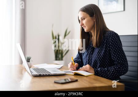 Frau macht sich Notizen, schaut sich Online-Webinare an, arbeitet an einem Projekt, das im Büro sitzt, schreibt sich Aufgaben und Ideen auf, Freiberuflerin, die mit einem Laptop arbeitet, um das Thema zu erkunden Stockfoto