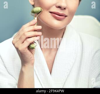 Face Jade Roller. Das verkürzte Gesicht einer erwachsenen Frau mit grünem Jaderoller für Anti-Aging-Verfahren Stockfoto