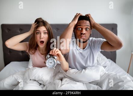 Schockiert multirassisches Paar mit Wecker, der zu spät zur Arbeit kommt, auf dem Bett sitzt und sich vor Angst den Kopf greift Stockfoto