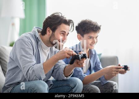 Gemeinsames Hobby. Überglücklich, Vater und Sohn spielen Videospiele und konkurrieren miteinander, sitzen auf dem Sofa Stockfoto