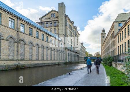 Zwei Personen gehen auf dem Treidelpfad des Leeds Liverpool Canal in Saltaire, West Yorkshire. Die Gebäude der Salts Mill säumen beide Seiten der Wasserstraße. Stockfoto