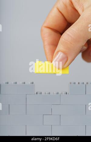 Ein gelber Ziegel legt eine Hand an eine graue Spielzeugwand. Das Konzept des Aufbaus, des Erreichens und der Vollendung des Ziels Stockfoto