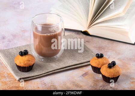 Muffins und ein Glas weißen Kaffee auf dem Tisch. Heiße Getränke, Kakao und offenes Papierbuch. Rustikales Stillleben im Vintage-Stil. Braune Cupcakes mit Beeren. Stockfoto