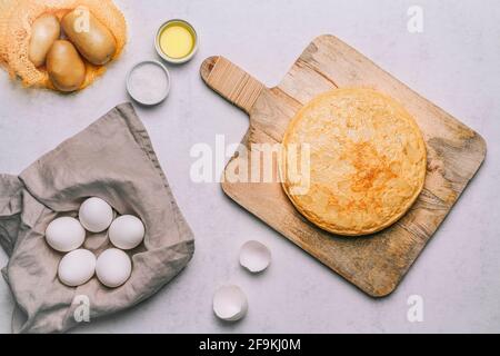 Luftaufnahme eines Kartoffelomeletts auf einem Retro-Holztisch, einem Korb mit weißen Eiern, Olivenöl und Salz und einem Orangennetz mit Kartoffeln auf weißem Marmor b Stockfoto