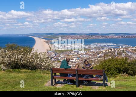 Familie auf der Bank mit Blick auf den Strand von Kesil Bank und Fortuneswell, Isle of Portland von Portland Heights, Portland, Weymouth Dorset, Großbritannien im April Stockfoto