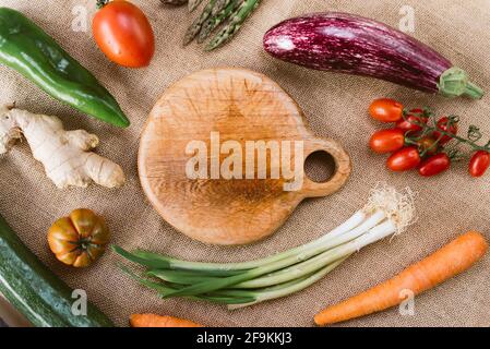 Draufsicht auf leeres Holzhackbrett, abgerundet vom frühlingshaften frischen Gemüse wie Tomaten und Karotten auf rustikalem Stoff. Konzept von Frühlingsnahrung und OR Stockfoto