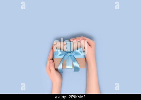 Weibliche Hände halten die Geschenkbox auf einem blauen pastellfarbenen Hintergrund. Geschenk gebunden mit einem Band. Festliches Konzept mit Copyspace. Stockfoto