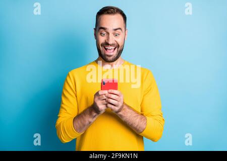 Foto-Portrait des staunenden Mannes in gelbem Outfit beim Lesen von Nachrichten Smartphone starrt lächelnd isoliert auf hellblauem Hintergrund Stockfoto