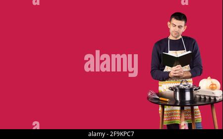 Serious hübscher junger Mann trägt professionelle Bäcker Schürze Kochen Rezept Buch lesen überrascht, lächelnd und Denken auf rotem Hintergrund mit leer Stockfoto