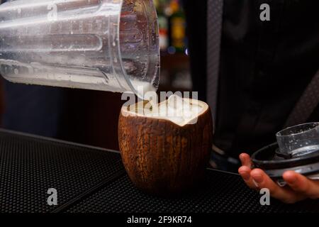 Der Barkeeper beendet die Zubereitung exotischer alkoholischer Cocktails in Kokosnuss, indem er eine Milch gießt. Nahaufnahme eines erfahrenen Barkeepers, der in der Bar einen Cocktail zubereitet. Stockfoto