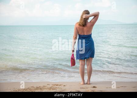 Eine Frau hält in der Hand eine Mesh-Einkaufstasche und steht am Rande des Meeres an einem Sandstrand. Ökologie-Konzept. Bewusster Konsum Stockfoto