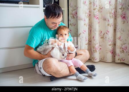 Entzückendes Baby sitzt in den Armen des Vaters und streichelt ein dekoratives Kaninchen. Vater zeigt kleines Kind osterhase. Haustiere in einer Familie mit Kindern Stockfoto