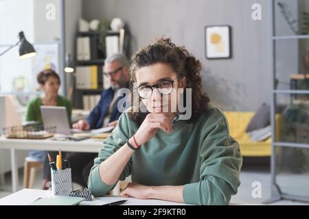 Porträt eines jungen Mitarbeiters in einer Brille, der die Kamera im Sitzen ansieht An seinem Tisch im Büro Stockfoto