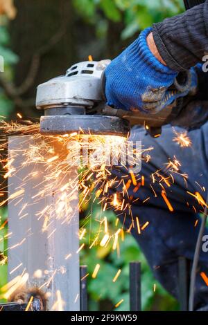 Die Hände des Baumeisters halten den Winkelschleifer fest, während Metall zerschnitten wird, und unter dem Schutzmantel des Winkelschleifers fliegen helle heiße Funken hervor. Stockfoto