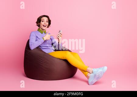 In voller Größe Profil Seite Foto von überrascht Mädchen sitzen Bohne Stuhl Punkt Finger Smartphone isoliert Pastellfarbe Hintergrund Stockfoto