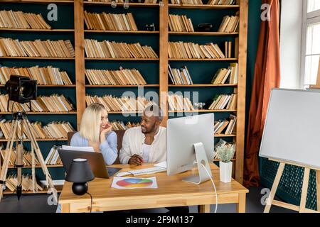 Glückliche, multiethnische Kollegen diskutieren über ein Projekt, sprechen über verschiedene Ideen, lächeln. Erfolgreiche Mitarbeiter, die ein Notebook verwenden und Papierkram machen Stockfoto